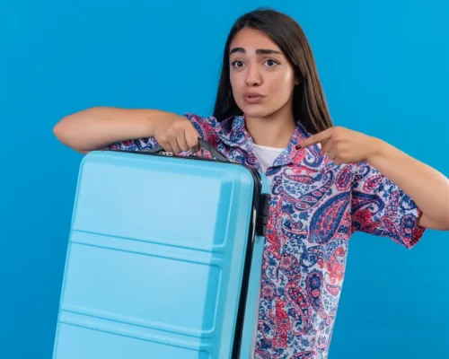 young-beautiful-traveler-woman-holding-blue-suitcase-pointing-with-finger-it-looking-confused-surprised-standing-blue-background young-beautiful-traveler-woman-holding-blue-suitcase-pointing-with-finger-it-looking-confused-surprised-standing-blue-background